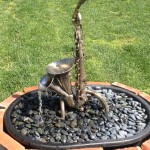 saxophone fountain, triple brass bells, saxophone body, water, river rocks, bricks, grass